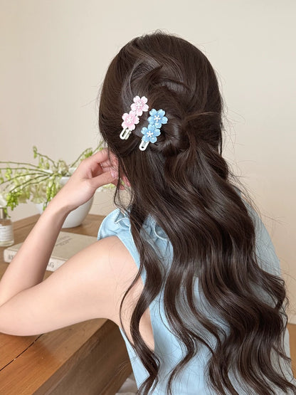A woman with long, dark brown wavy hair is shown from the back, wearing two small flower-shaped hair clips in pastel pink and light blue. She is wearing a light blue sleeveless top and sitting at a wooden table in a softly lit indoor setting.