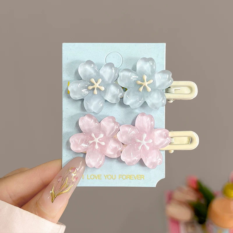 A hand holds a light blue card displaying two sets of flower-shaped hair clips. The top set is translucent light blue with cream centers, and the bottom set is translucent pink with cream centers.