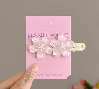 A pair of translucent pink cherry blossom hair clips attached to a pink display card, held by a hand with floral nail art against a neutral gray background.