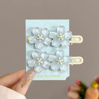 Four translucent light blue flower-shaped hair clips with cream centers, attached to a light blue display card held by a hand with decorated nails, against a soft taupe background.