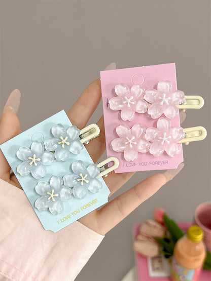 A hand holds two flower-shaped hair clips on display cards. One card is light blue with matching pale blue flowers and gold text reading 'I LOVE YOU FOREVER.