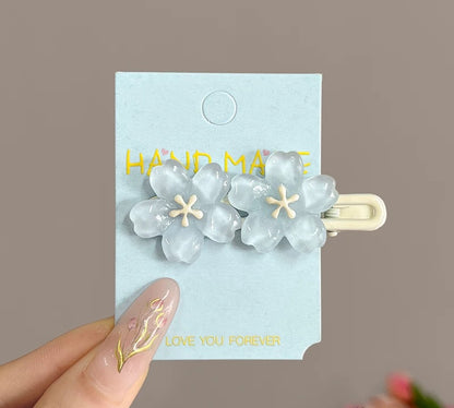 A pair of translucent pale blue flower hair clips with cream centers, attached to a light blue display card held by a hand with gold nail art against a soft taupe background.