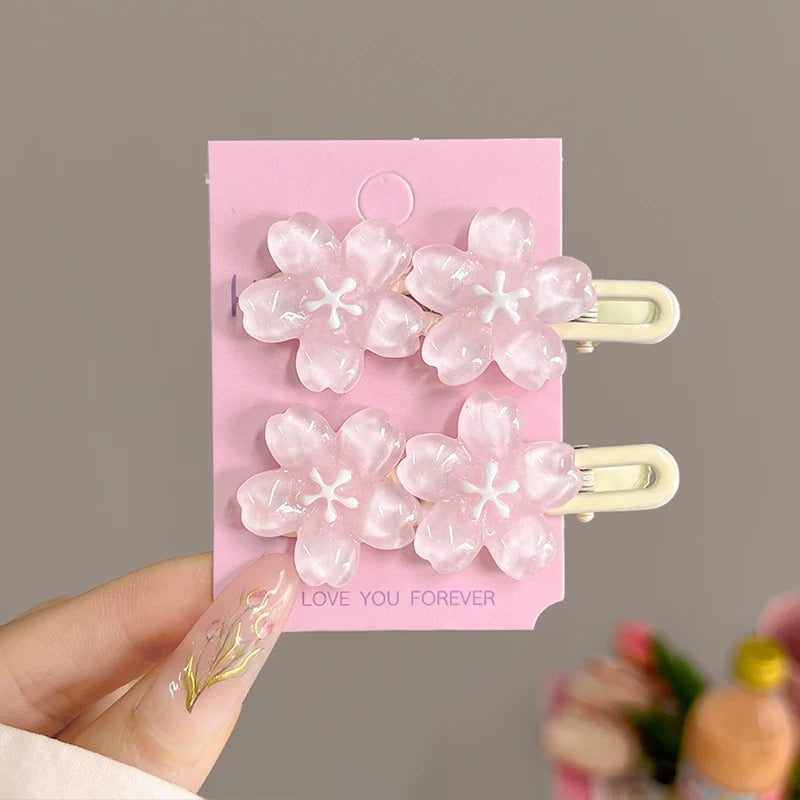 A hand holds a pink display card featuring four translucent pink cherry blossom hair clips with cream-colored metal clasps. The card has 'LOVE YOU FOREVER' printed on it, and the background is a soft, blurred gray.
