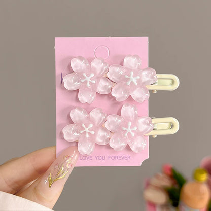 A hand holds a pink display card featuring four translucent pink cherry blossom hair clips with cream-colored metal clasps. The card has 'LOVE YOU FOREVER' printed on it, and the background is a soft, blurred gray.