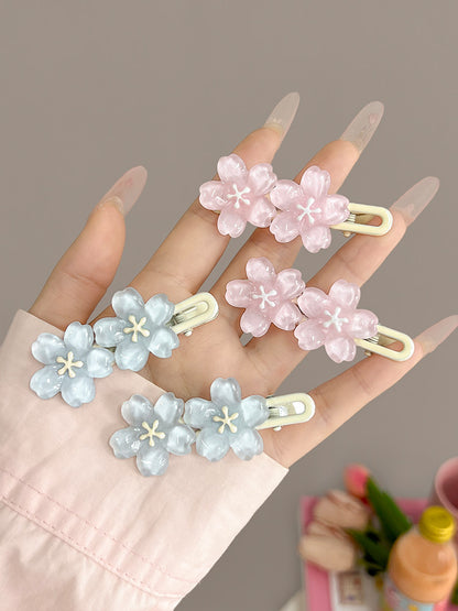 Four flower-shaped hair clips in pastel pink and light blue, held in a hand with long clear acrylic nails against a soft taupe background.