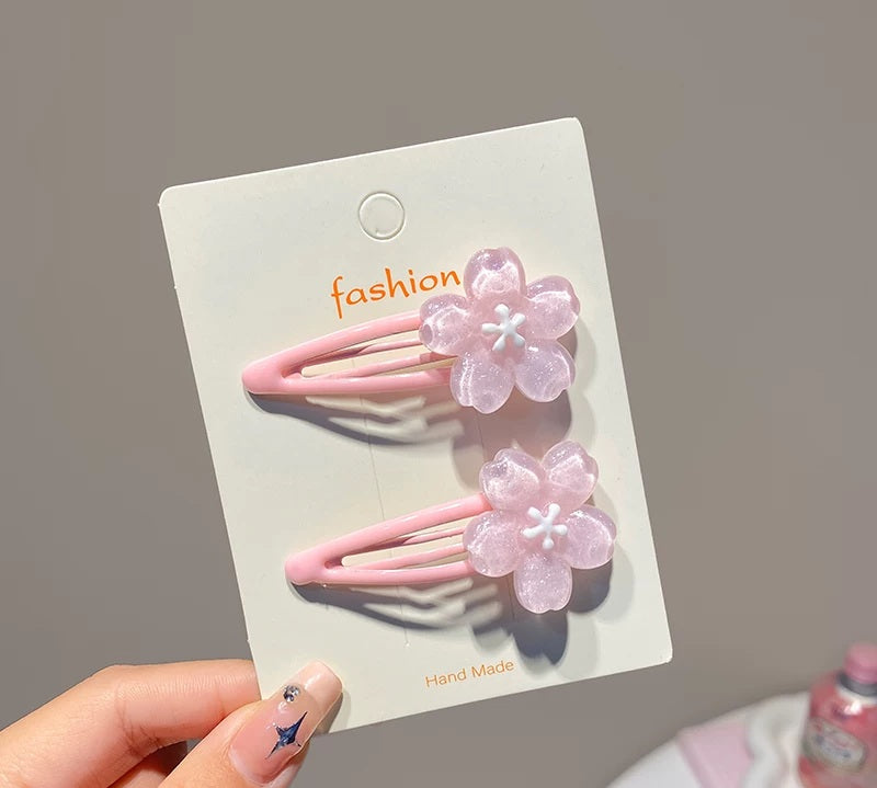 A hand holds a white card displaying two pink hair clips shaped like translucent pink cherry blossoms, with the word 'fashion' printed on the card against a soft gray background.