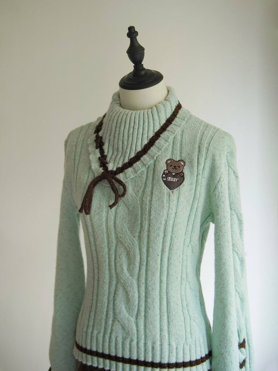 A light mint green cable-knit sweater displayed on a mannequin torso. The sweater features a dark brown braided tie detail at the collar, a brown embroidered bear patch on the left chest, and dark brown trim at the hem.