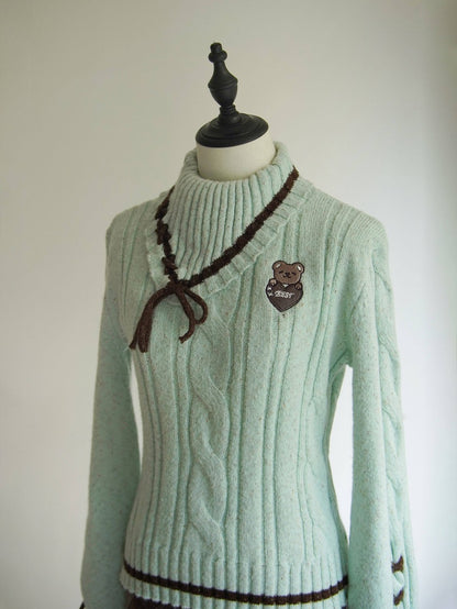 A light mint green cable-knit sweater displayed on a mannequin torso. The sweater features a dark brown braided tie detail at the collar, a brown embroidered bear patch on the left chest, and dark brown trim at the hem.