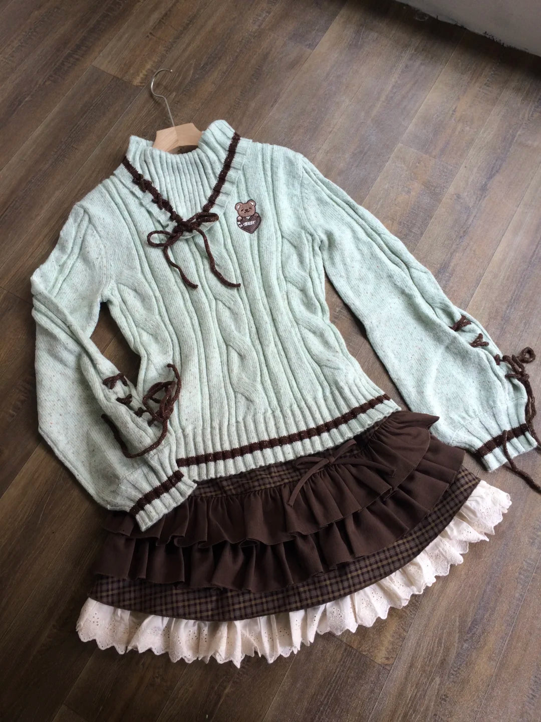 A light sage green cable-knit sweater dress with dark brown lace-up details, a ruffled brown waistband, and a layered ruffle skirt with a cream ruffle trim, laid flat on a wooden floor.
