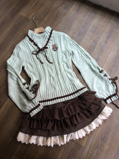 A light sage green cable-knit sweater dress with dark brown lace-up details, a ruffled brown waistband, and a layered ruffle skirt with a cream ruffle trim, laid flat on a wooden floor.