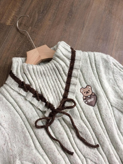 A light beige ribbed knit turtleneck sweater with dark brown trim and a matching bow, featuring a small embroidered teddy bear holding a heart patch on the chest, displayed on a wooden hanger over a dark wood floor.
