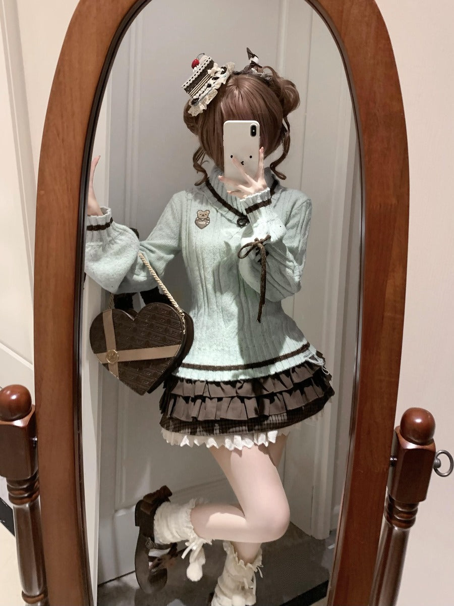 A person taking a mirror selfie wearing a light mint green long-sleeve sweater with dark brown lace trim, a matching dark brown ruffled pleated mini skirt with white lace trim, white thigh-high stockings, and brown lace-up boots. They are holding a brown heart-shaped crossbody bag.