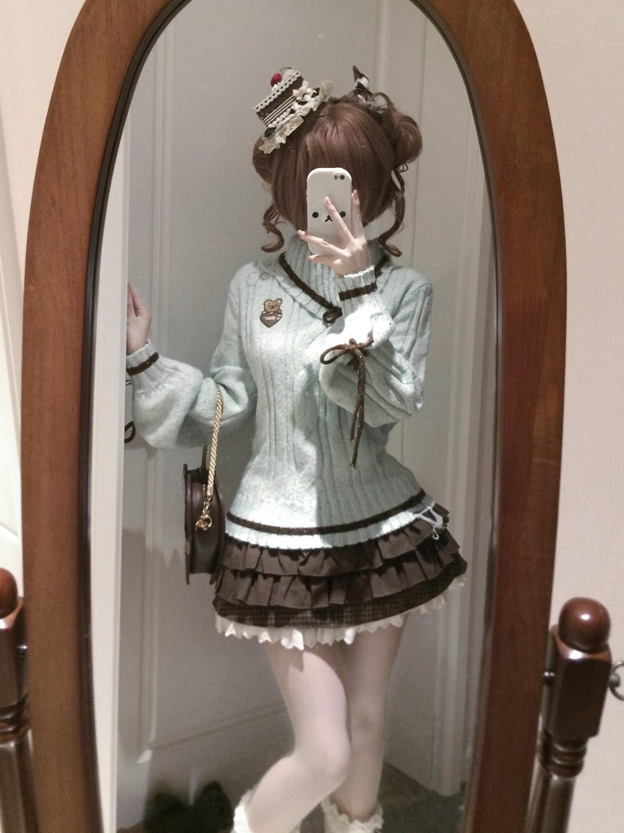 A person taking a mirror selfie wearing a light blue knit sweater dress with dark brown ruffled trim and a matching dark brown pleated skirt, paired with a decorative headpiece and carrying a small black handbag.