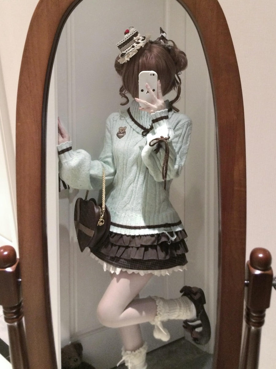A person taking a mirror selfie wearing a light mint green knit sweater with black trim, a dark brown pleated mini skirt, white thigh-high stockings, and a small brown crossbody bag. The photo is taken in front of an arched wooden mirror.