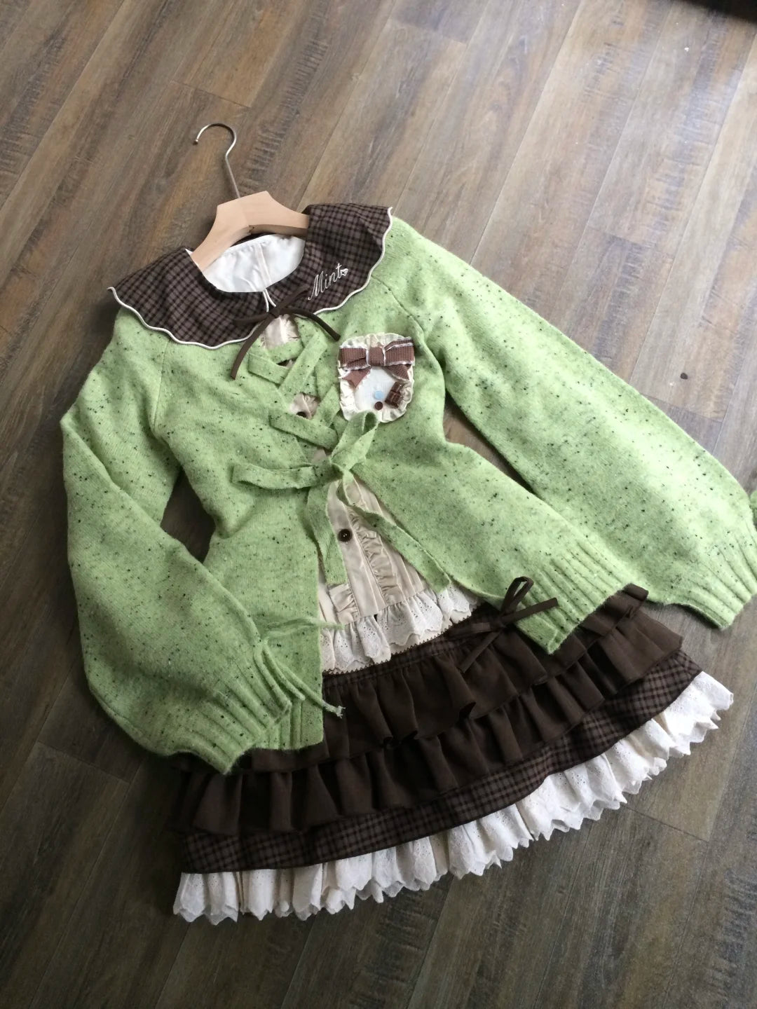 A green knit cardigan with brown plaid collar and cuffs, layered over a cream ruffled blouse and a brown ruffled skirt with white lace trim, displayed on a wooden hanger over a dark wood floor.