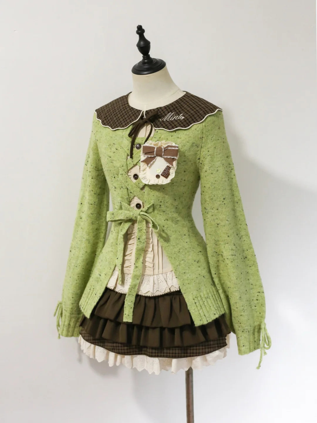 A light green knit cardigan sweater with a brown plaid collar, featuring a decorative embroidered pocket with a bow and the word 'Mia' on the chest, displayed on a mannequin against a plain white background.