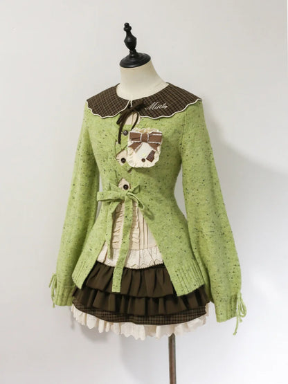 A light green knit cardigan sweater with a brown plaid collar, featuring a decorative embroidered pocket with a bow and the word 'Mia' on the chest, displayed on a mannequin against a plain white background.