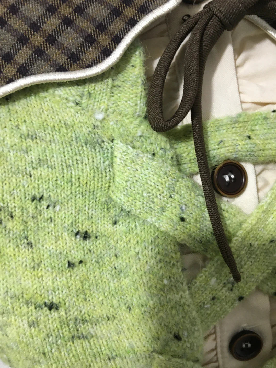 Close-up of a knit cardigan or sweater detail showing a light green heathered knit body, dark brown drawstring, cream lining, and a brown-and-black plaid collar.