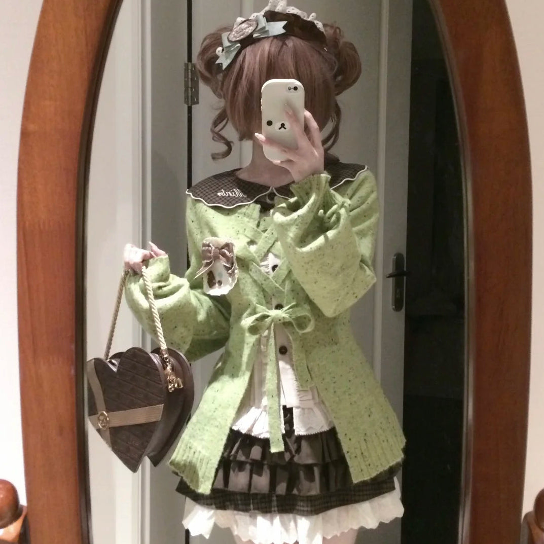 A person taking a mirror selfie wearing a light green lace-up cardigan over a white and black ruffled Lolita-style dress, holding a brown heart-shaped handbag.