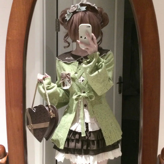 A person taking a mirror selfie wearing a light green lace-up cardigan over a white and black ruffled Lolita-style dress, holding a brown heart-shaped handbag.