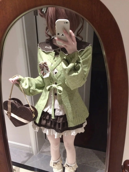 A person taking a mirror selfie wearing a light green knit cardigan with dark brown lace trim, a white and dark brown pleated dress, white thigh-high stockings, and holding a brown heart-shaped handbag.