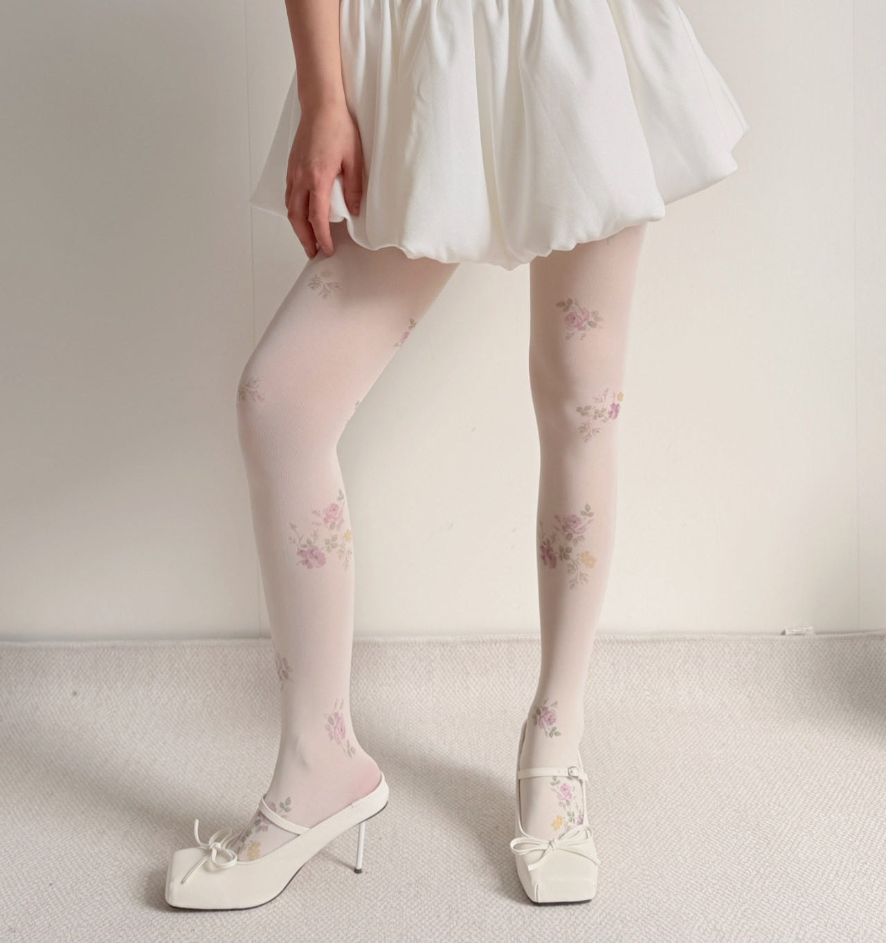 A person wearing sheer white tights with a delicate pastel floral print, styled with a white mini skirt and white Mary Jane heels against a neutral cream background.