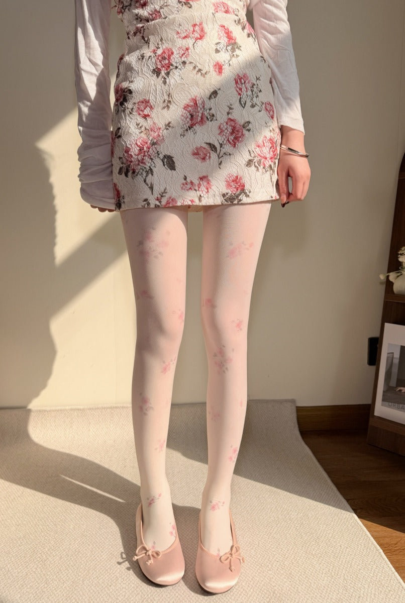 A model wearing a white long-sleeve top, a white floral mini skirt, and sheer light pink floral tights with matching pink bow flats, standing indoors on a light-colored rug.