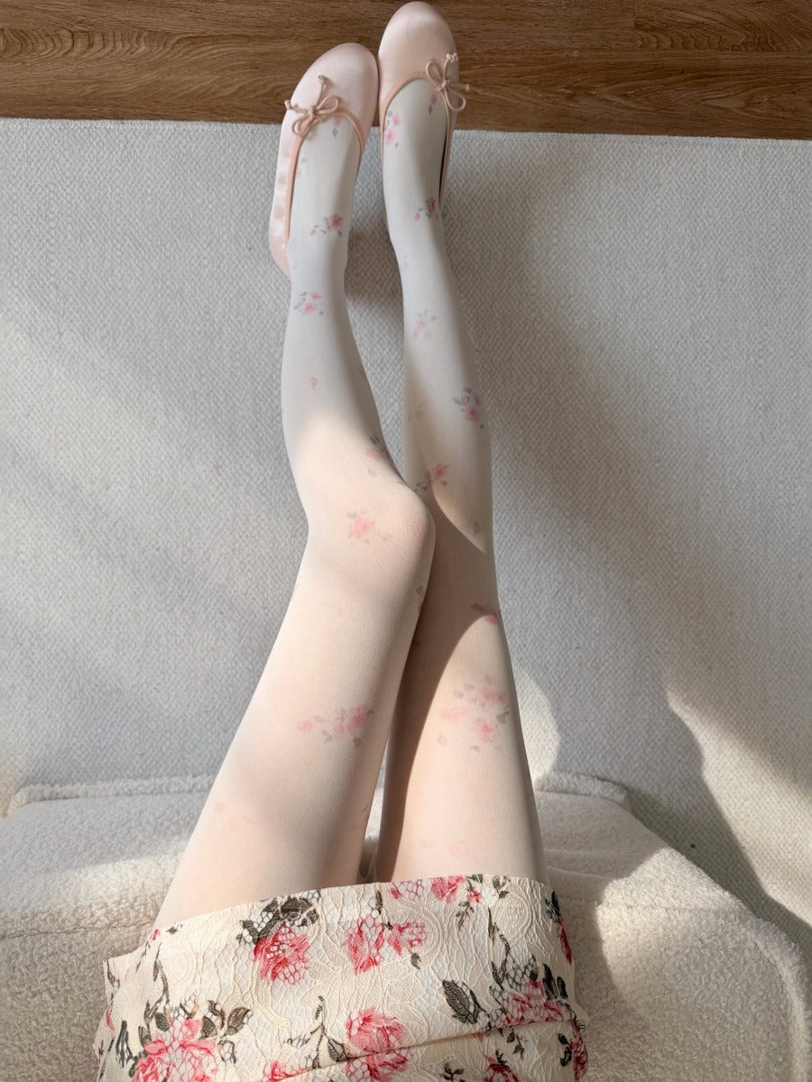 A person seated indoors wearing sheer white tights with a delicate pink floral print and matching pointed flats with bows. The floral hem of the skirt or dress is also visible, and the scene is lit by natural sunlight with soft shadows on a light textured surface.