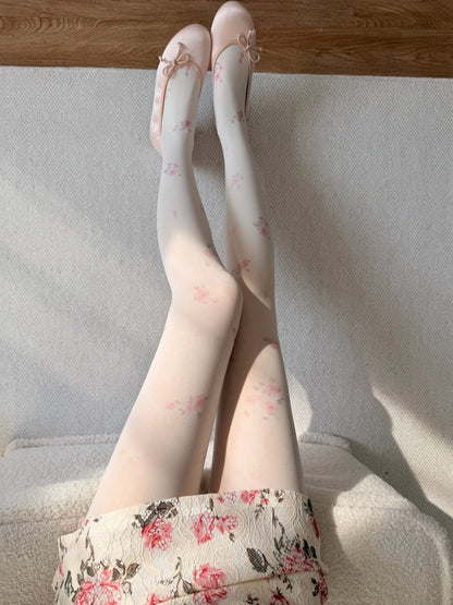 A person seated indoors wearing sheer white tights with a delicate pink floral print and matching pointed flats with bows. The floral hem of the skirt or dress is also visible, and the scene is lit by natural sunlight with soft shadows on a light textured surface.