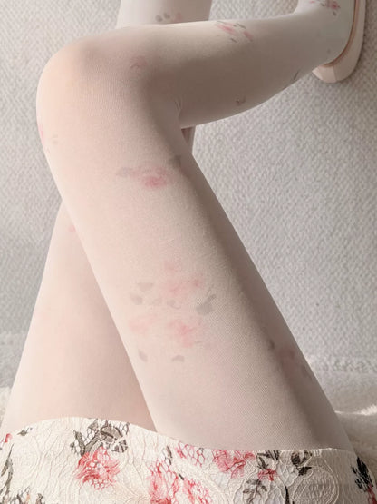 A close-up of a person's legs wearing sheer white tights with a delicate pink and gray floral pattern, paired with a matching floral skirt against a textured light gray background.