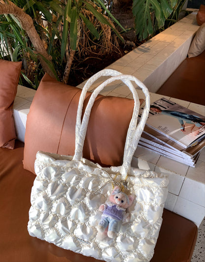 A white quilted fabric tote bag with pearl-like trim and a small plush character charm, resting on a brown leather bench in an indoor setting with plants and magazines in the background.