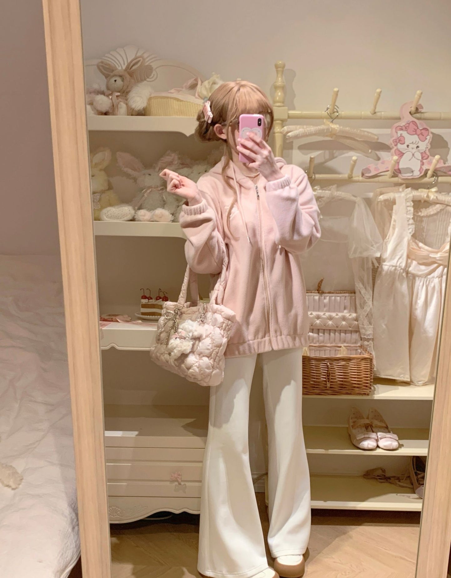 A person taking a mirror selfie in a bedroom showing a light pink knit zip-up hoodie, white wide-leg pants, and a clear tote bag with white pom-poms.