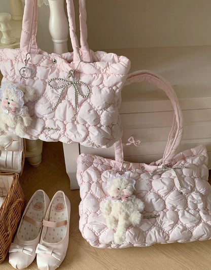 Two light pink quilted shoulder bags with rhinestone bow details and plush teddy bear charms, displayed on a wooden floor next to white ballet flats.
