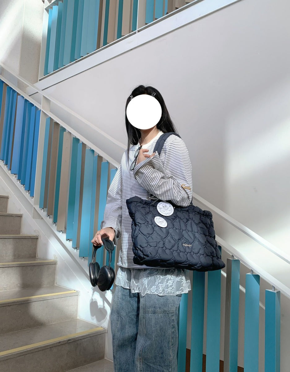 A person standing on an indoor staircase holding a black quilted shoulder bag with two round white tags attached, wearing a striped long-sleeve top and blue jeans.