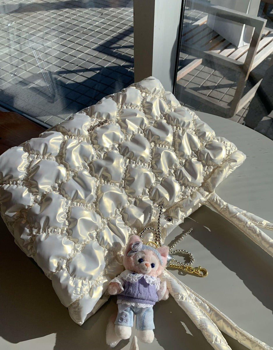 A cream-colored quilted puffer handbag with a matching shoulder strap, styled on a table with a small plush pig keychain.