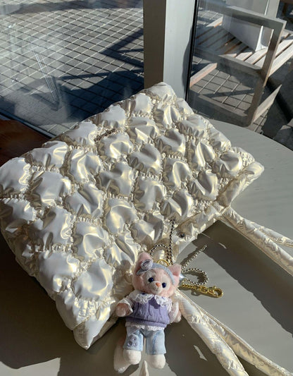 A cream-colored quilted puffer handbag with a matching shoulder strap, styled on a table with a small plush pig keychain.