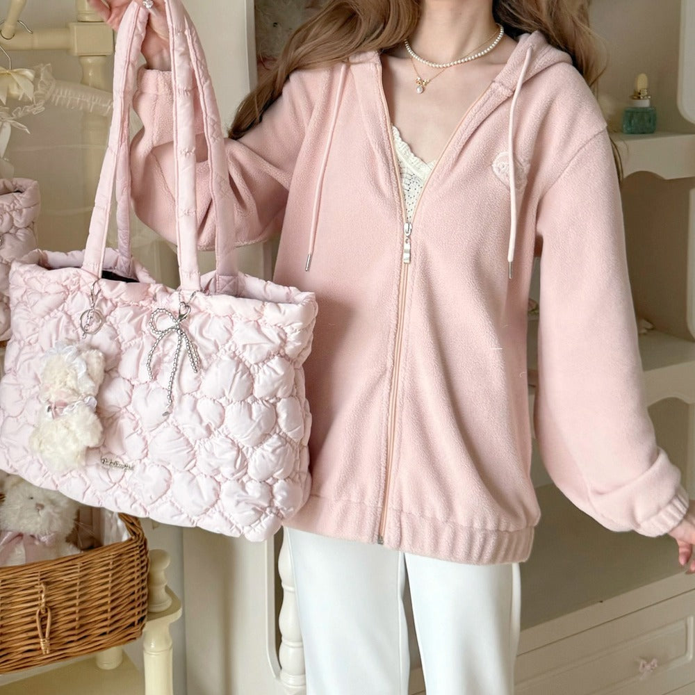 A woman wearing a light pink zip-up hoodie and white pants, holding a matching pink quilted tote bag with a bow and rhinestone charm in a soft indoor setting.