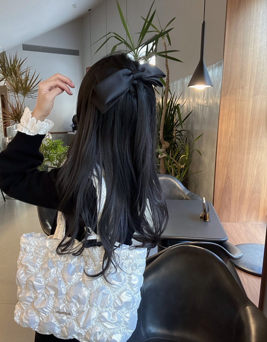 A person with long black hair seen from behind, wearing a large black bow headband and a black-and-white outfit, standing in a modern indoor setting with plants and furniture visible.