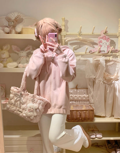 A person taking a mirror selfie in a room decorated with pastel items, wearing a light pink zip-up hoodie, white pants, and carrying a large pink plush teddy bear shoulder bag.