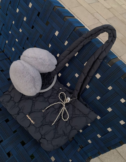 A dark gray quilted tote bag with a pearl-bead bow and a small gold logo, styled on a blue woven outdoor bench next to fluffy light gray earmuffs.