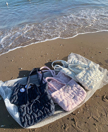 Three quilted puffer tote bags in black, light pink, and white are arranged on a plastic sheet on a sandy beach with the ocean waves in the background.