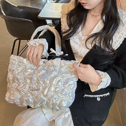 A person in a black cardigan and cream ruffled blouse holds a large iridescent white ruched shoulder bag with pearl-like trim in a cafe setting.
