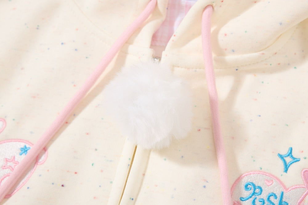 Close-up of a cream-colored hoodie with pink drawstrings, a fluffy white pom-pom, and pastel embroidered details.