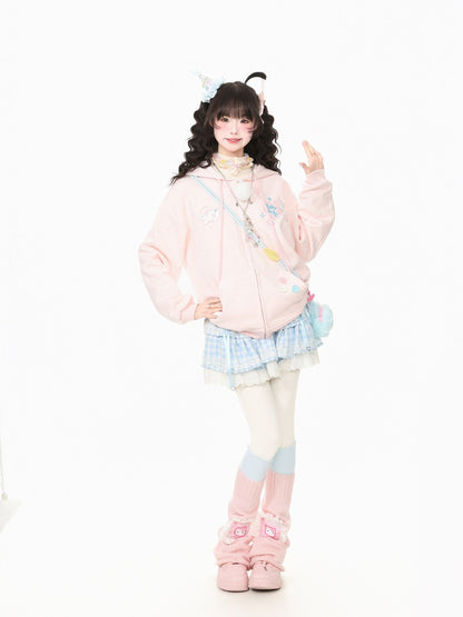 A young woman modeling a pastel pink zip-up hoodie with cute cartoon graphics, paired with a light blue and white ruffled pleated skirt, white tights with pink and blue striped socks, and pink fuzzy boots, set against a plain white studio background.