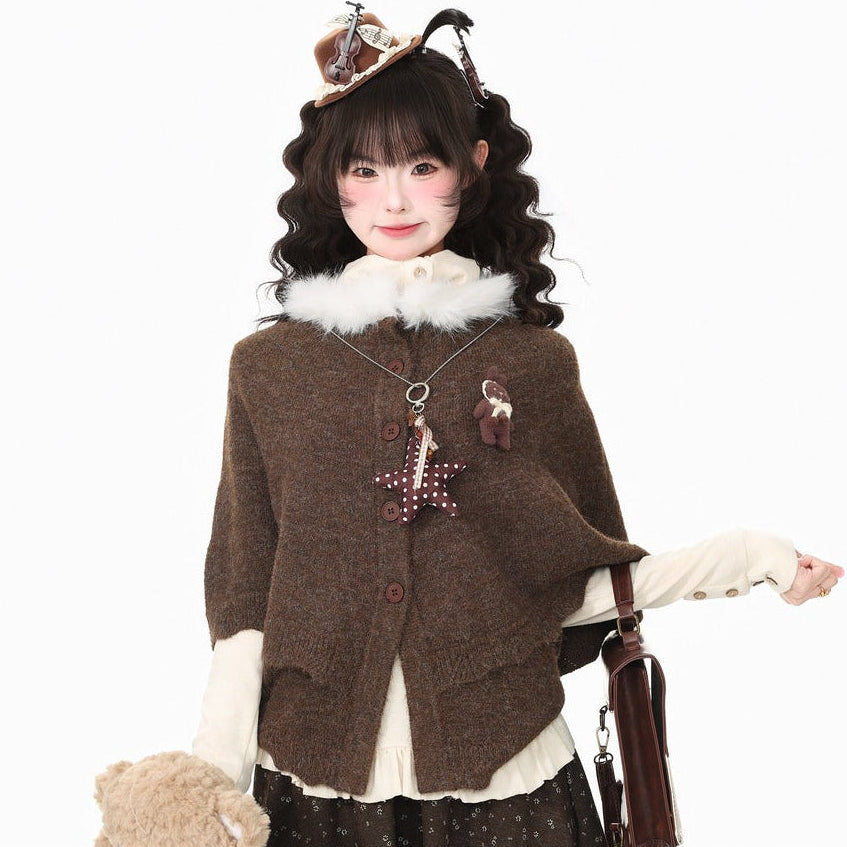 A model wearing a cozy brown knit poncho cardigan with a fluffy white collar, styled with a dark patterned skirt, a brown teddy bear accessory, and a small hat, set against a plain white background.