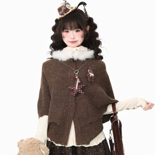 A model wearing a cozy brown knit poncho cardigan with a fluffy white collar, styled with a dark patterned skirt, a brown teddy bear accessory, and a small hat, set against a plain white background.