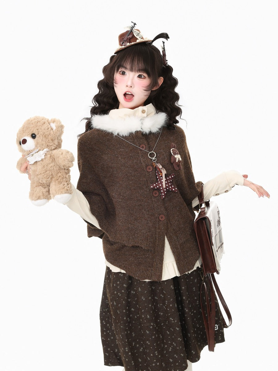 A young woman modeling a brown knit cardigan with a white faux-fur collar, styled with a dark patterned skirt, cream sleeves, a brown teddy bear plush, and a small hat against a plain white background.