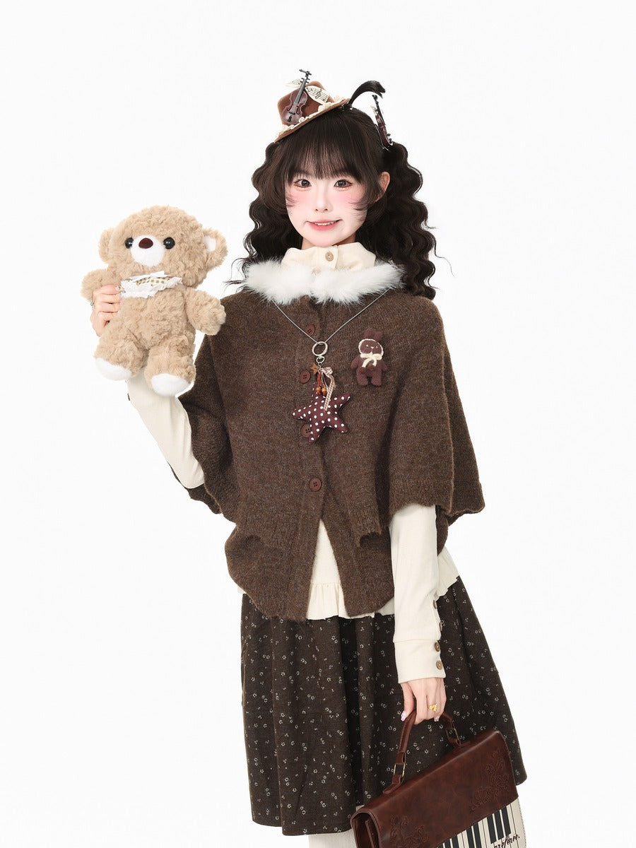 A young girl models a cozy brown knit cardigan with a white faux-fur collar, styled with a brown patterned skirt, cream long-sleeve shirt, and a plush teddy bear accessory, against a solid white background.