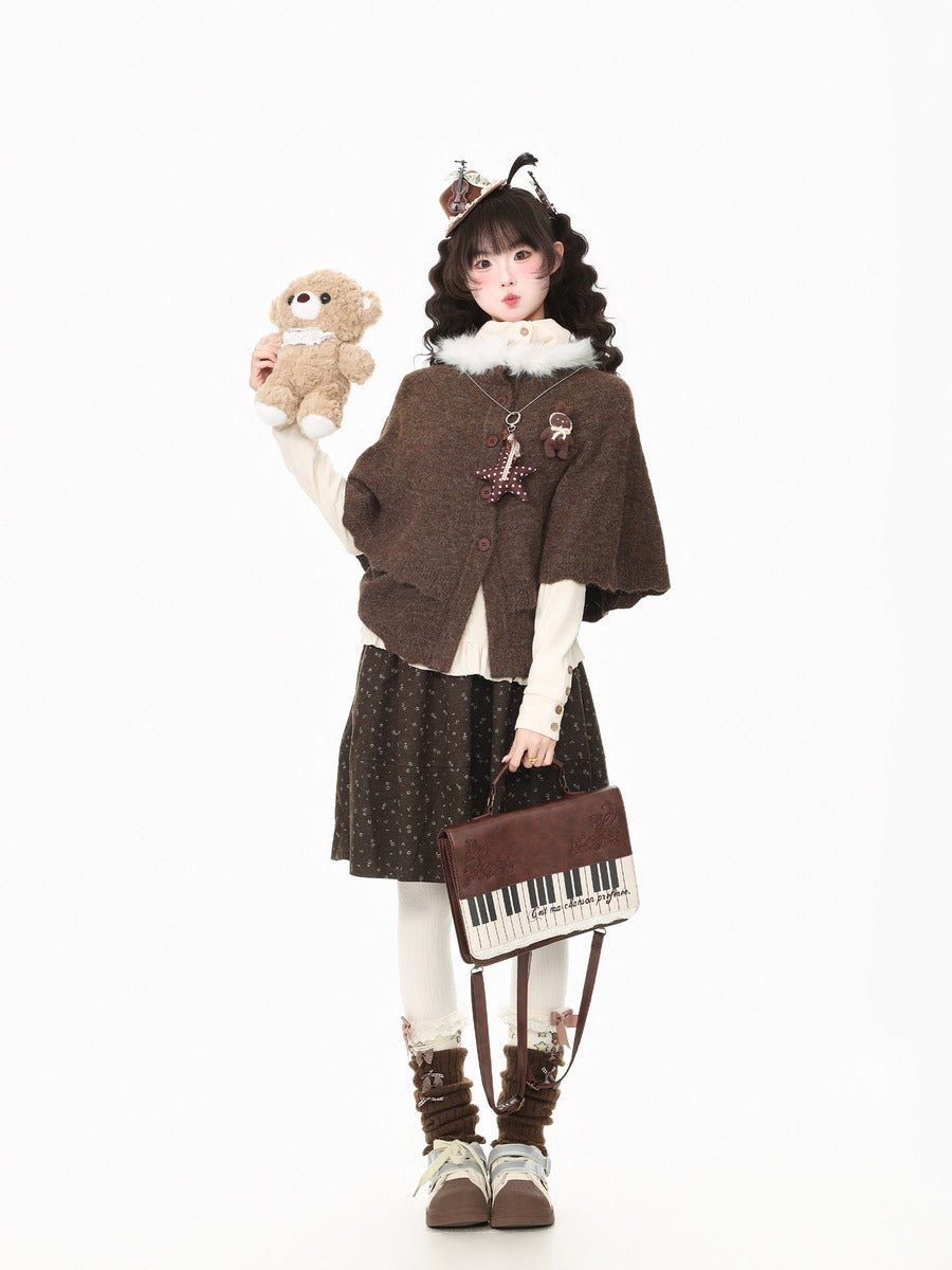 A young girl models a cozy, gothic-style outfit consisting of a brown knit cape, a dark patterned skirt, white tights, and brown lace-up boots. She holds a small teddy bear and a mini piano-shaped shoulder bag against a solid white background.
