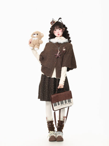 A young girl models a cozy, gothic-style outfit consisting of a brown knit cape, a dark patterned skirt, white tights, and brown lace-up boots. She holds a small teddy bear and a mini piano-shaped shoulder bag against a solid white background.